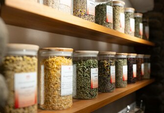 Several glass containers with loose teas and herbs are arranged on wooden shelves in a tea area.