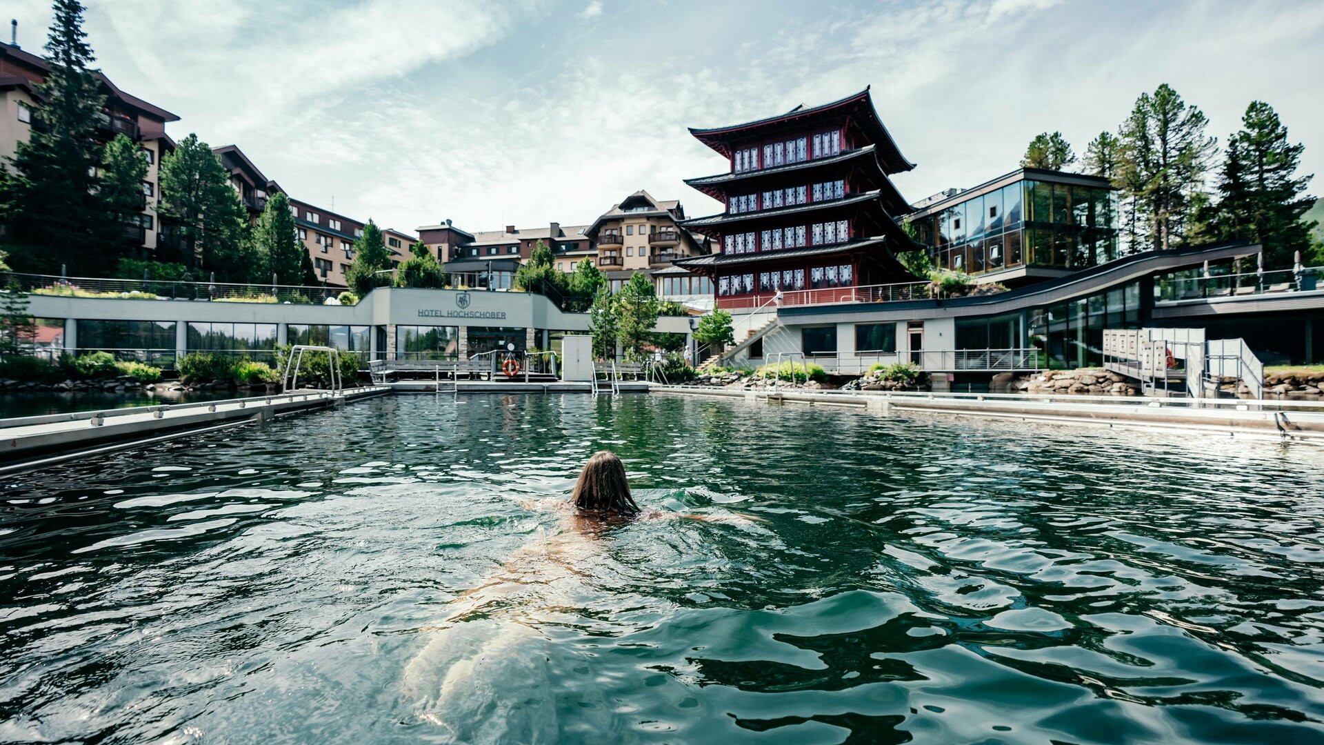 A person swims in the heated lake pool with the Hotel Hochschober buildings and lakeside area in the background.