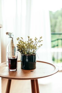 Ein kleiner Holztisch mit einer Karaffe, einem Glas Rotwein und einer Blumenvase steht vor einem hellen Fenster.