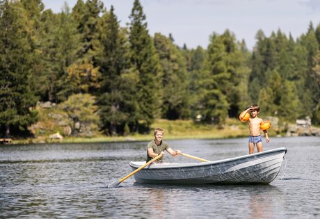 Zwei Kinder sind in einem Ruderboot auf dem See unterwegs, eines rudert, das andere trägt Schwimmflügel.