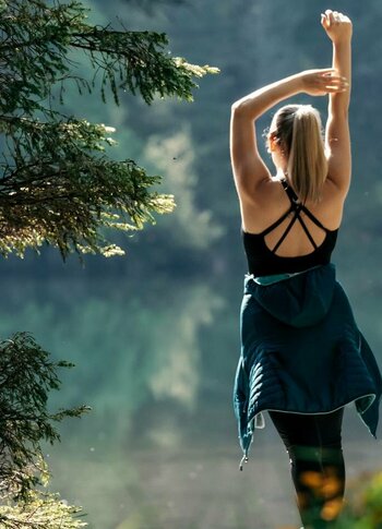Urlaubsangebote für das ganze Jahr - Turracher Höhe Eine Person steht mit erhobenen Armen auf einem Weg, umgeben von Bäumen und bunten Wildblumen. Im Hintergrund spiegelt sich ein ruhiger See in der Sonne. Die Szene strahlt Ruhe und Naturverbundenheit aus.