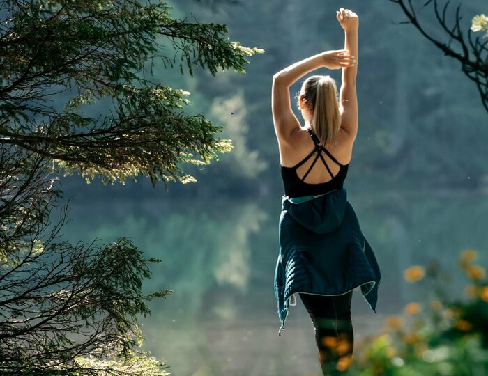 Urlaubsangebote für das ganze Jahr - Turracher Höhe Eine Person steht mit erhobenen Armen auf einem Weg, umgeben von Bäumen und bunten Wildblumen. Im Hintergrund spiegelt sich ein ruhiger See in der Sonne. Die Szene strahlt Ruhe und Naturverbundenheit aus.