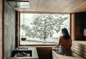 Eine Frau sitzt in der Holzsauna und blickt durch das große Fenster auf die verschneite Landschaft.