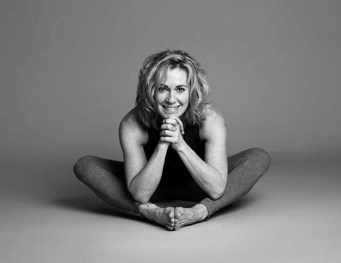 A woman sits in a yoga pose on the floor and smiles at the camera.