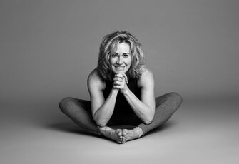 A woman sits in a yoga pose on the floor and smiles at the camera.
