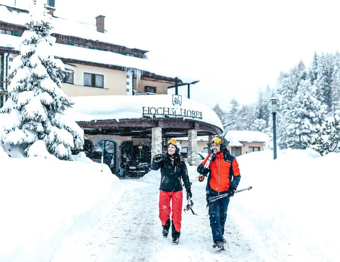 Ski holiday at Hotel Hochschober in snow Two skiers walk with skis and helmets along a snow-covered path in front of Hotel Hochschober.