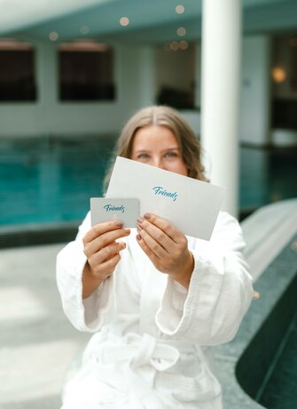 A woman in a white bathrobe holds up a card and an envelope in front of an indoor pool.