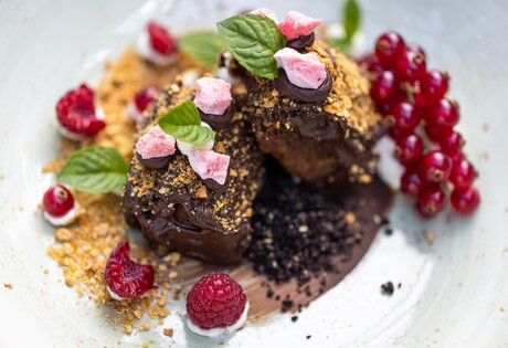 Zwei Schokoladendesserts mit Beeren, Krokant und Minze angerichtet auf einem hellen Teller.