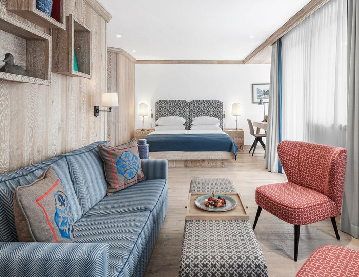 Ein helles Hotelzimmer mit Sofa, Sessel, kleinem Tisch und Bett vor bodentiefen Vorhängen.