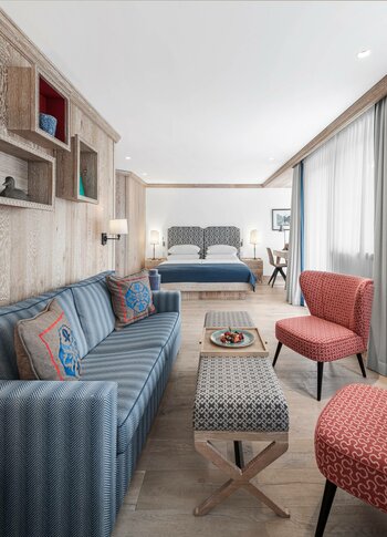 A bright hotel room with a sofa, armchair, small table and bed beside floor-length curtains.