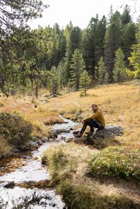 Ein Mann sitzt auf einem Felsen an einem kleinen Bach in einer herbstlichen Wald- und Alm-Landschaft.