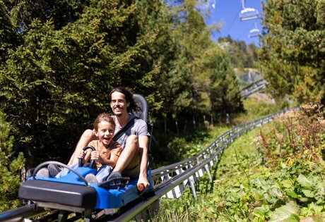 Ein Mann und ein Kind fahren gemeinsam auf einer Sommerrodelbahn durch eine grüne Berglandschaft.