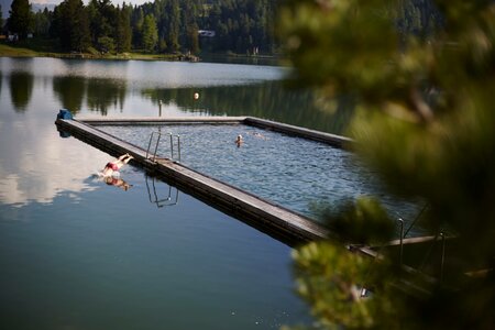 Ein schwimmendes See-Bad mit Holzsteg liegt ruhig im Turracher See, umgeben von Wald und Berglandschaft.