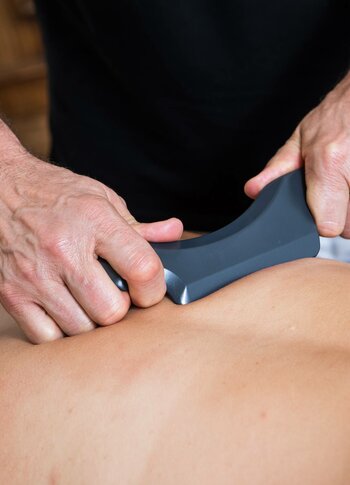 Eine Masseurin bearbeitet den Rücken einer liegenden Person mit einem Massagegerät.