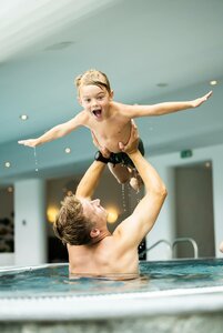 Ein Mann hebt ein Kind lachend im Innenpool hoch, beide mit ausgebreiteten Armen.