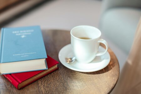 Auf einem kleinen Holztisch stehen eine Tasse Kaffee, ein Unterteller und mehrere Bücher.