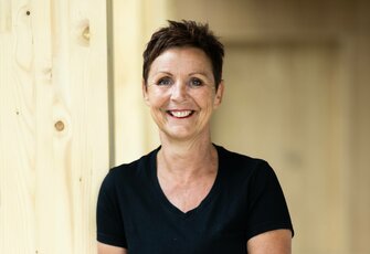 Eine lächelnde Frau steht vor einer hellen Holzwand und trägt ein schwarzes Shirt.