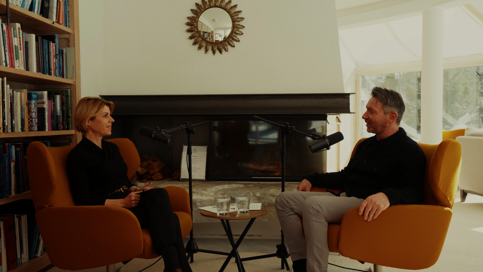 A woman and a man sit in armchairs by the fireplace, speaking into podcast microphones.