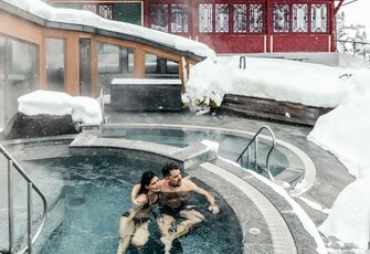 A couple sits in the heated lake pool while snow falls around the pool edge.