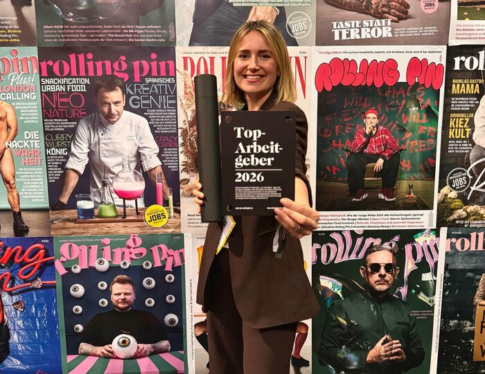 Eine Frau hält vor einer Wand mit Rolling-Pin-Magazincovern ein schwarzes Schild mit der Aufschrift „Top-Arbeitgeber 2026“.