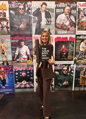 Rolling Pin Magazin mit Frau vor Coverwand Eine Frau hält vor einer Wand mit Rolling-Pin-Magazincovern ein schwarzes Schild mit der Aufschrift „Top-Arbeitgeber 2026“.