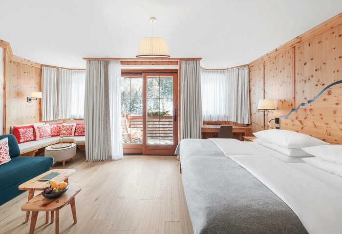 Das Deluxe Zirbenzimmer zeigt ein großes Bett, eine Sitzgruppe mit Sofa und einen hellen Balkonbereich mit Holzverkleidung.