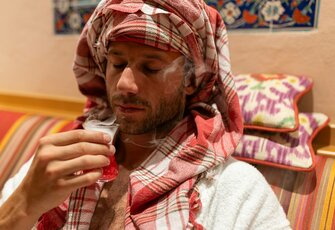 A man in a towel turban smells a small glass of tea or water in the oriental hammam.