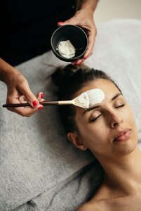Eine Frau erhält im Spa eine Gesichtsbehandlung, während Creme mit einem Pinsel auf die Stirn aufgetragen wird.