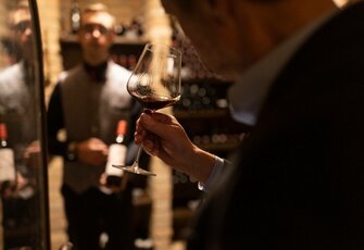 Eine Hand hält ein Weinglas mit Rotwein; im Hintergrund steht ein Kellner mit Flasche in einem warm beleuchteten Hotelbereich.