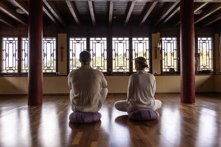Zwei Personen sitzen im Yoga-Raum auf Meditationskissen vor den großen Fenstern.