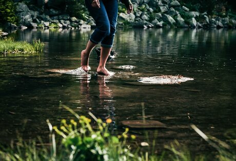 Eine Person watet barfuß durch flaches Wasser am Seeufer, im Hintergrund liegen Steine und Ufervegetation.